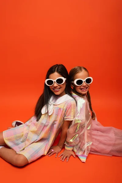 Веселые и модные девушки в солнечных очках сидят и улыбаются в камеру на оранжевом фоне — стоковое фото