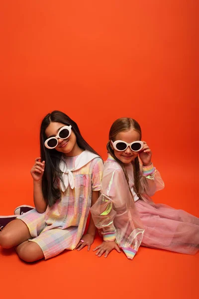 Lächelnde Mädchen in trendiger Kleidung und Sonnenbrille, die in die Kamera schauen, während sie auf orangefarbenem Hintergrund sitzen — Stockfoto