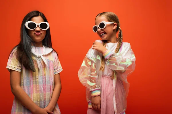 Rire fille en veste de pluie transparente et des lunettes de soleil en regardant grimacing ami isolé sur orange — Photo de stock