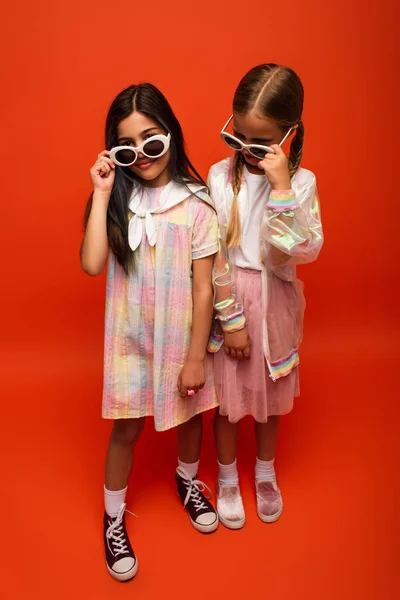 Full length of preteen girls in dress and rain jacket holding sunglasses on orange background — Stock Photo