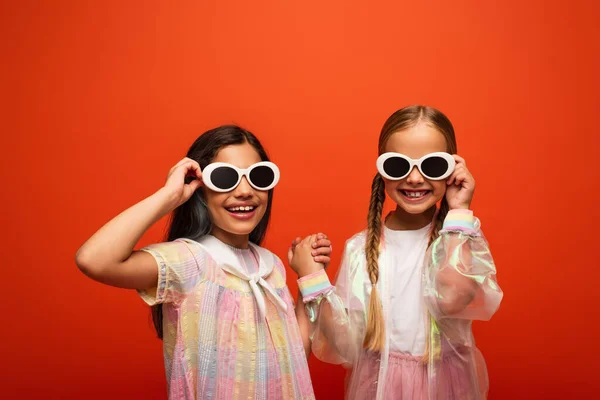 Joyful girls holding hands and touching trendy sunglasses isolated on orange — Stock Photo