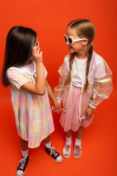 Vue grand angle des filles en vêtements à la mode et lunettes de soleil se tenant la main et se souriant sur fond orange — Photo de stock