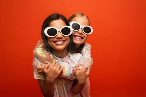 Ragazze felici in eleganti occhiali da sole che abbracciano e sorridono isolati su arancione — Foto stock