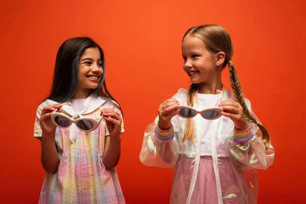 Gaies filles en robe et veste de pluie transparente tenant des lunettes de soleil et se regardant isolés sur orange — Photo de stock