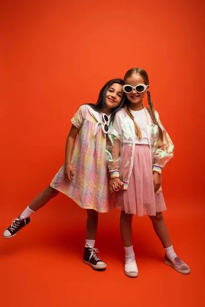 Full length of happy girl in sunglasses holding hands with friend while posing on orange background — Stock Photo