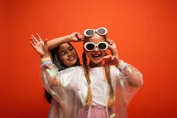 Aufgeregtes Mädchen mit zwei Sonnenbrillen, die Hand winkend, während sie Spaß mit einem Freund hat, der isoliert auf orange steht — Stockfoto