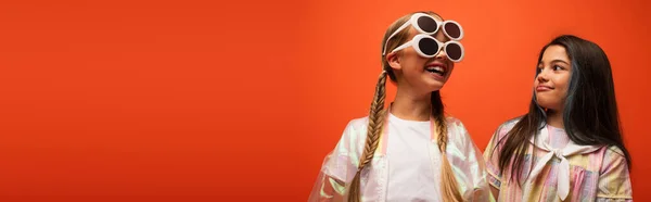 Excitada chica en dos gafas de sol divirtiéndose cerca morena amigo aislado en naranja, pancarta - foto de stock