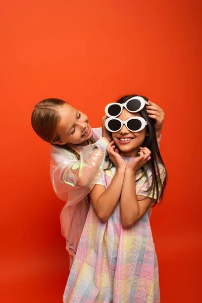 Smiling girl putting two sunglasses on smiling friend isolated on orange — Stock Photo