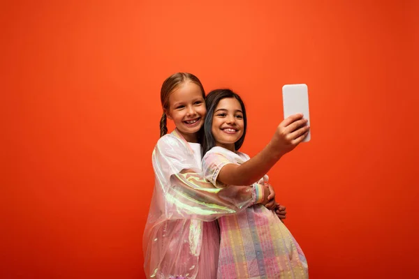 Girl in rain jacket hugging friend in dress taking selfie on cellphone isolated on orange — Stock Photo