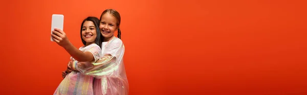 Брюнетка дівчина бере селфі з другом у дощовій куртці, обіймаючи її ізольовано на помаранчевому, банер — стокове фото