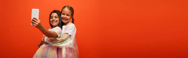 Brünettes Mädchen macht Selfie mit Freundin in Regenjacke und umarmt sie isoliert auf orangefarbenem Banner — Stockfoto