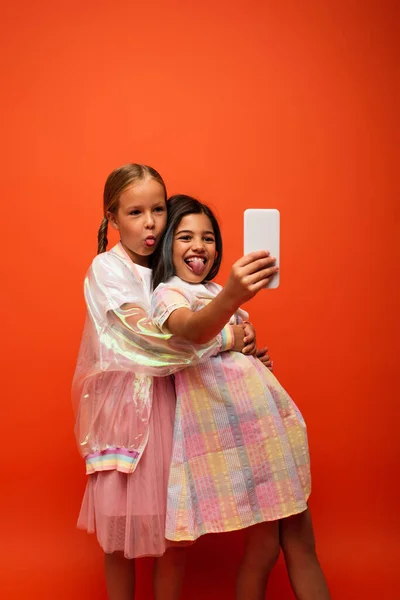 Alegre preadolescente niñas sobresaliendo lenguas mientras toma selfie en el teléfono móvil aislado en naranja - foto de stock