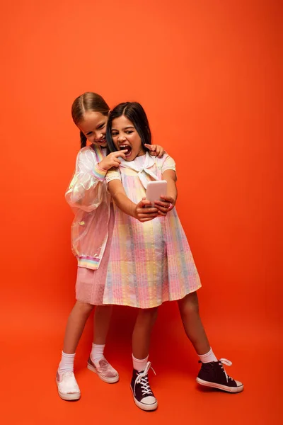 Lächelndes Kind zeigt aufgeregtes Mädchen mit offenem Mund an, während es Spaß hat und ein Selfie auf orangefarbenem Hintergrund macht — Stockfoto