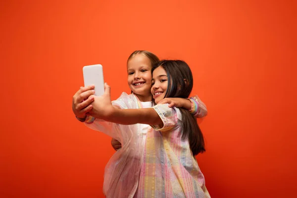 Niños alegres en ropa de moda abrazando y tomando selfie en el teléfono inteligente aislado en naranja - foto de stock