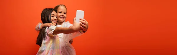 Souriant préadolescentes dans des vêtements élégants étreignant et prenant selfie sur smartphone isolé sur orange, bannière — Photo de stock