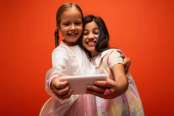 Excited friends hugging and taking selfie on mobile phone on blurred foreground isolated on orange — Stock Photo