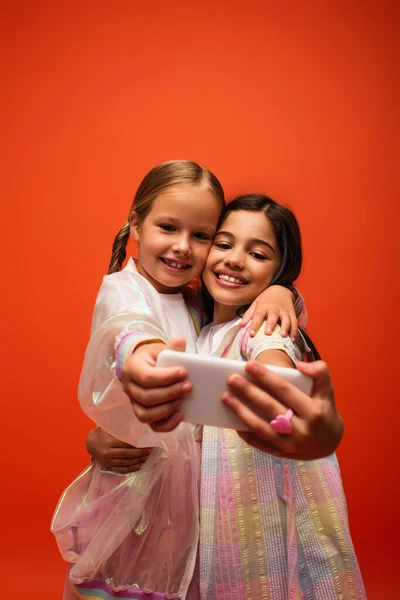 Веселые девочки-подростки в модной одежде обнимаются, делая селфи на смартфоне, изолированном на оранжевом — стоковое фото