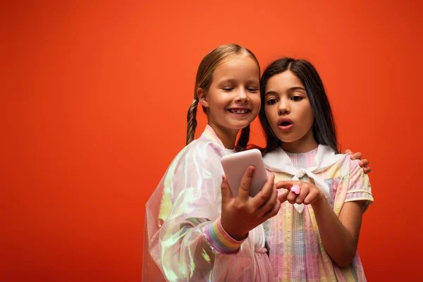Здивована дівчина вказує на мобільний телефон біля усміхненого друга ізольовано на помаранчевому — Stock Photo