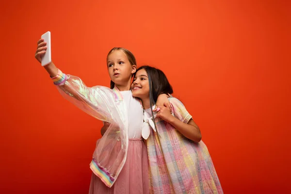 Girl in transparent rain jacket taking selfie with happy brunette friend isolated on orange — Stock Photo