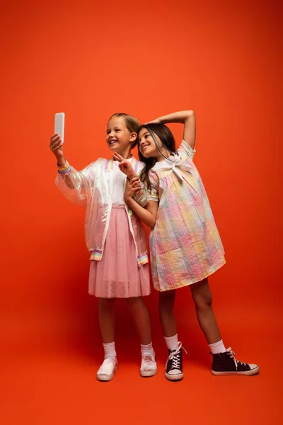 Chica feliz mostrando signo de victoria cerca de amigo tomando selfie en el teléfono móvil en fondo naranja - foto de stock