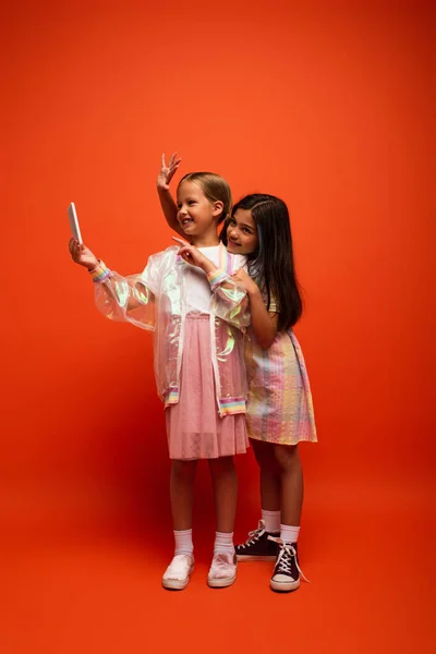 Повна довжина щасливих дівчат, що махають рукою і вказують пальцем, беручи селфі на помаранчевий фон — стокове фото