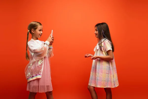 Vista lateral da menina morena gestos e falando perto de amigo gravação de vídeo no smartphone isolado em laranja — Fotografia de Stock