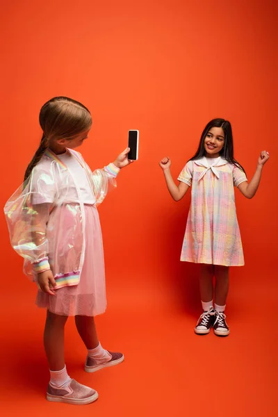 Comprimento total da menina feliz mostrando gesto de vitória perto de amigo tirar foto no celular no fundo laranja — Fotografia de Stock