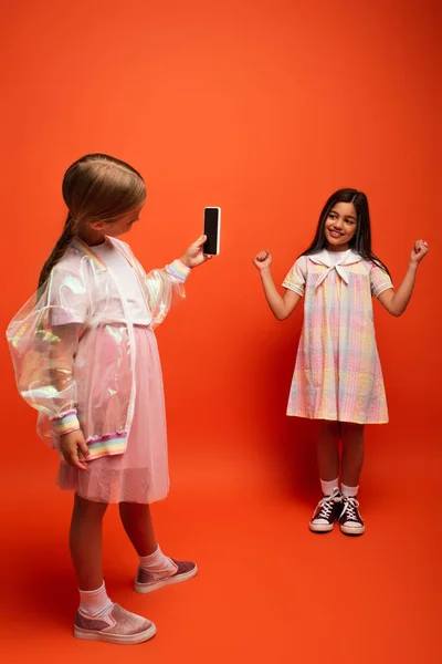 Full length of happy girl showing win gesture near friend taking photo on mobile phone on orange background — Stock Photo