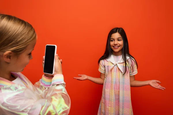 Girl with mobile phone taking photo of smiling brunette friend posing with open arms isolated on orange — Stock Photo
