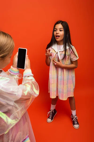 Chica sorprendida hablando y señalando con el dedo cerca de amigo grabación de vídeo en el teléfono inteligente sobre fondo naranja - foto de stock