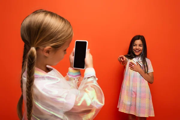 Girl with pigtails taking photo of laughing friend pointing with finger isolated on orange — Stock Photo