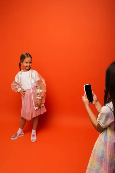Chica sonriente en chaqueta de lluvia transparente posando con la mano en la cadera cerca de amigo tomando fotos en el teléfono móvil sobre fondo naranja - foto de stock