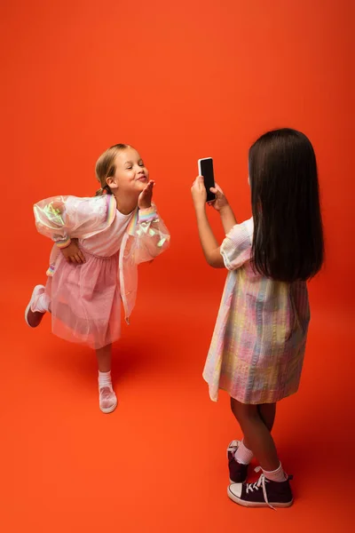 Alegre chica posando con la mano en la cadera y soplando aire beso cerca amigo tomando foto en el teléfono celular en naranja fondo - foto de stock