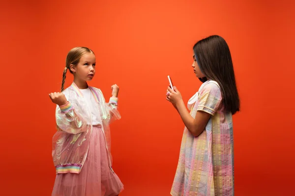 Brünettes Mädchen mit Handy fotografiert nachdenklichen Freund in Regenjacke isoliert auf orange — Stockfoto