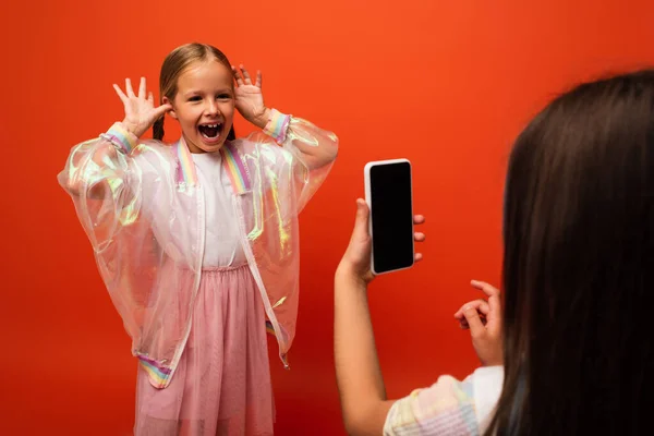 Fille drôle avec bouche ouverte gesticulant ami proche prendre des photos sur smartphone isolé sur orange — Photo de stock