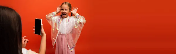 Дитина фотографує дивовижну дівчину з відкритим ротом жестикулюючи руками біля голови ізольовано на помаранчевому, банер — стокове фото