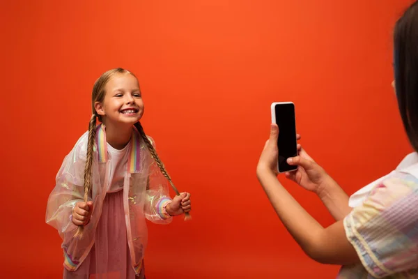 Menina engraçada segurando suas tranças perto amigo tirar foto no smartphone isolado em laranja — Fotografia de Stock