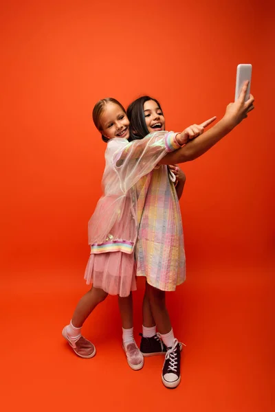 Девочка в полный рост показывает пальцем рядом с другом, делающим селфи на мобильном телефоне на оранжевом фоне — стоковое фото