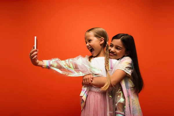 Chica sonriente abrazando amigo emocionado con la boca abierta tomar selfie en el teléfono inteligente aislado en naranja - foto de stock