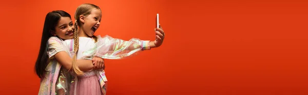 Ragazza sorridente abbracciando amico eccitato con la bocca aperta prendendo selfie su smartphone isolato su arancione, banner — Foto stock