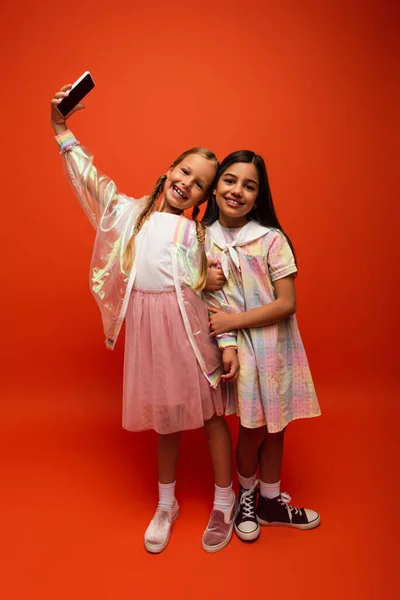 Longitud completa de chica feliz en impermeable tomando selfie con amigo en vestido sobre fondo naranja - foto de stock