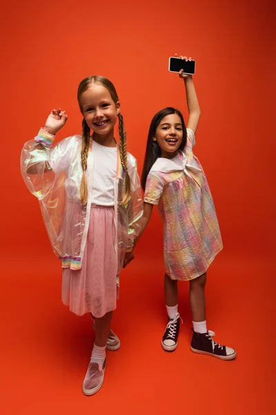 Excited girl showing smartphone with blank screen while holding hands with happy friend on orange background — Stock Photo