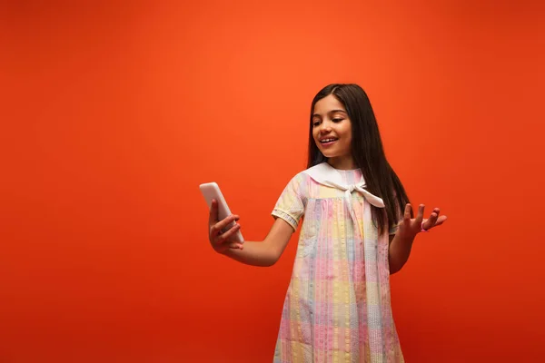 Alegre morena chica en vestido tomando selfie en el teléfono celular y apuntando con la mano aislada en naranja - foto de stock