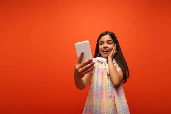Вражена і щаслива дівчина торкається щоки і приймає селфі на смартфон ізольовано на помаранчевому — стокове фото