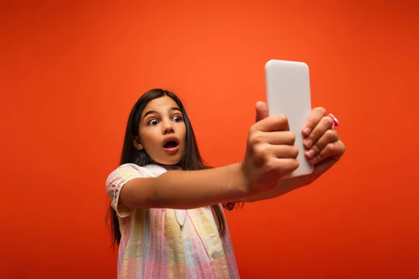 Brunette girl with shocked face expression taking selfie on smartphone isolated on orange — Stock Photo
