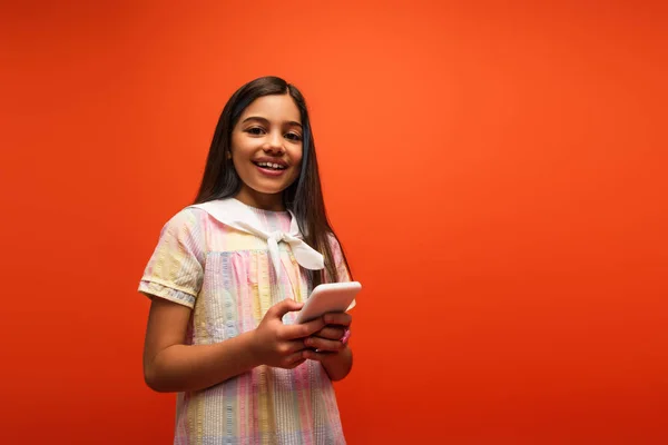 Morena feliz chica en vestido sosteniendo el teléfono móvil y mirando a la cámara aislada en naranja - foto de stock