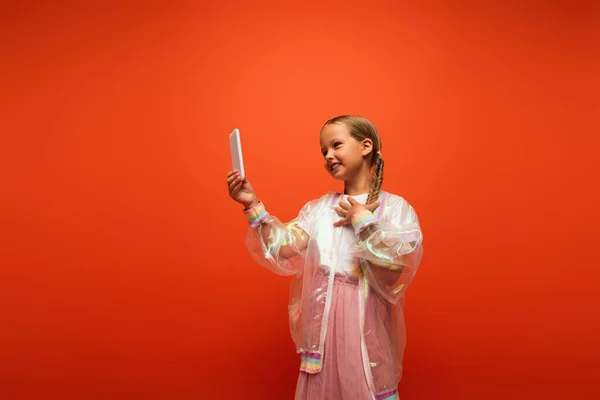Pleased girl in trendy rain jacket touching chest while talking during video call isolated on orange — Stock Photo