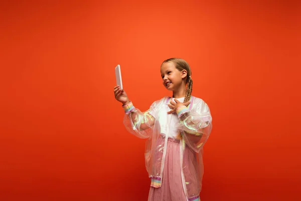 Heureux fille à la mode veste de pluie toucher la poitrine tout en parlant lors de l'appel vidéo isolé sur orange — Photo de stock