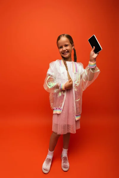 Longitud completa de chica alegre tocando la coleta y mostrando el teléfono inteligente con pantalla en blanco sobre fondo naranja - foto de stock