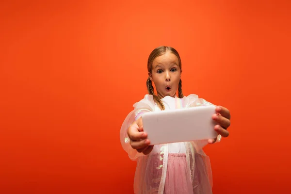 Chica con la boca abierta y la expresión de la cara emocionada tomar selfie en el teléfono celular aislado en naranja - foto de stock
